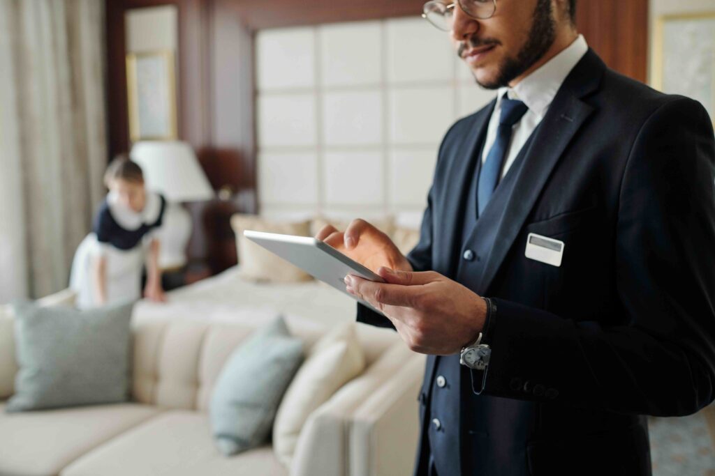 Hotel manager on tablet PMS in hotel room as housekeeper cleans in background